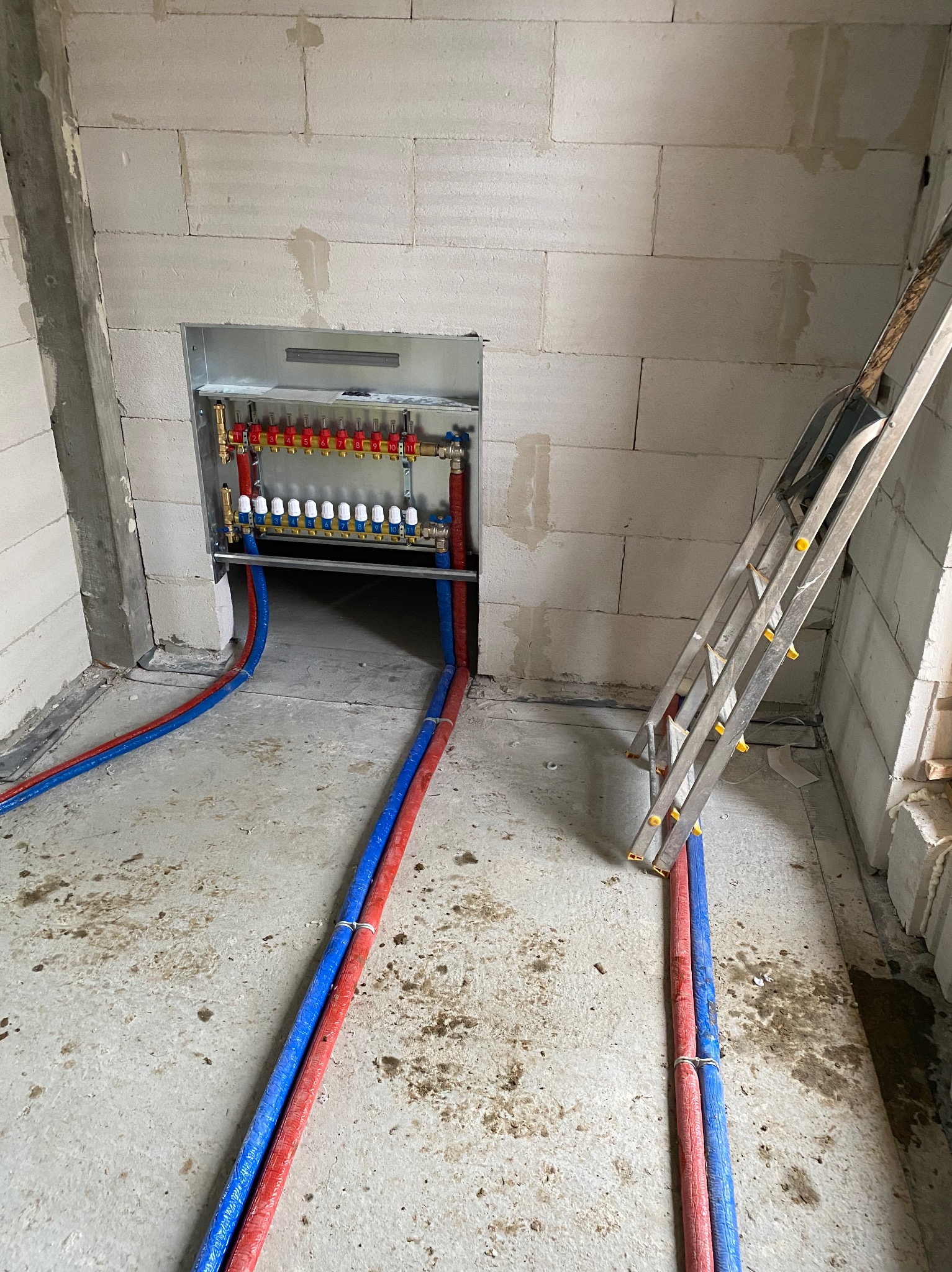 Rozdzielacz ogrzewania podłogowego z czerwono-niebieskimi rurami w surowym wnętrzu, drabina oparta o ścianę z betonu komórkowego.