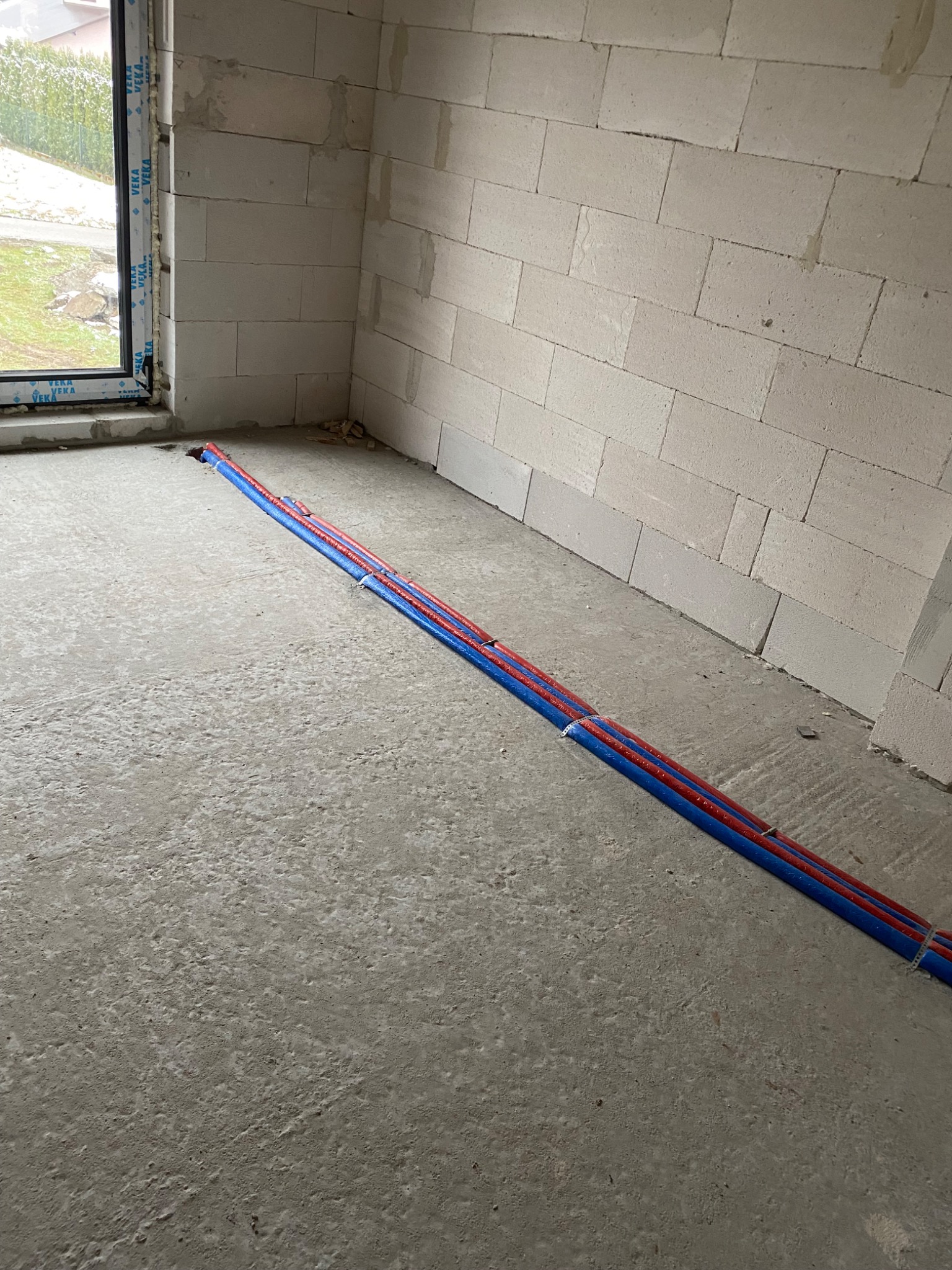 Czerwono-niebieskie rury instalacji grzewczej ułożone na surowym betonowym podłożu w nowo budowanym pomieszczeniu z białymi ścianami z betonu komórkowego.