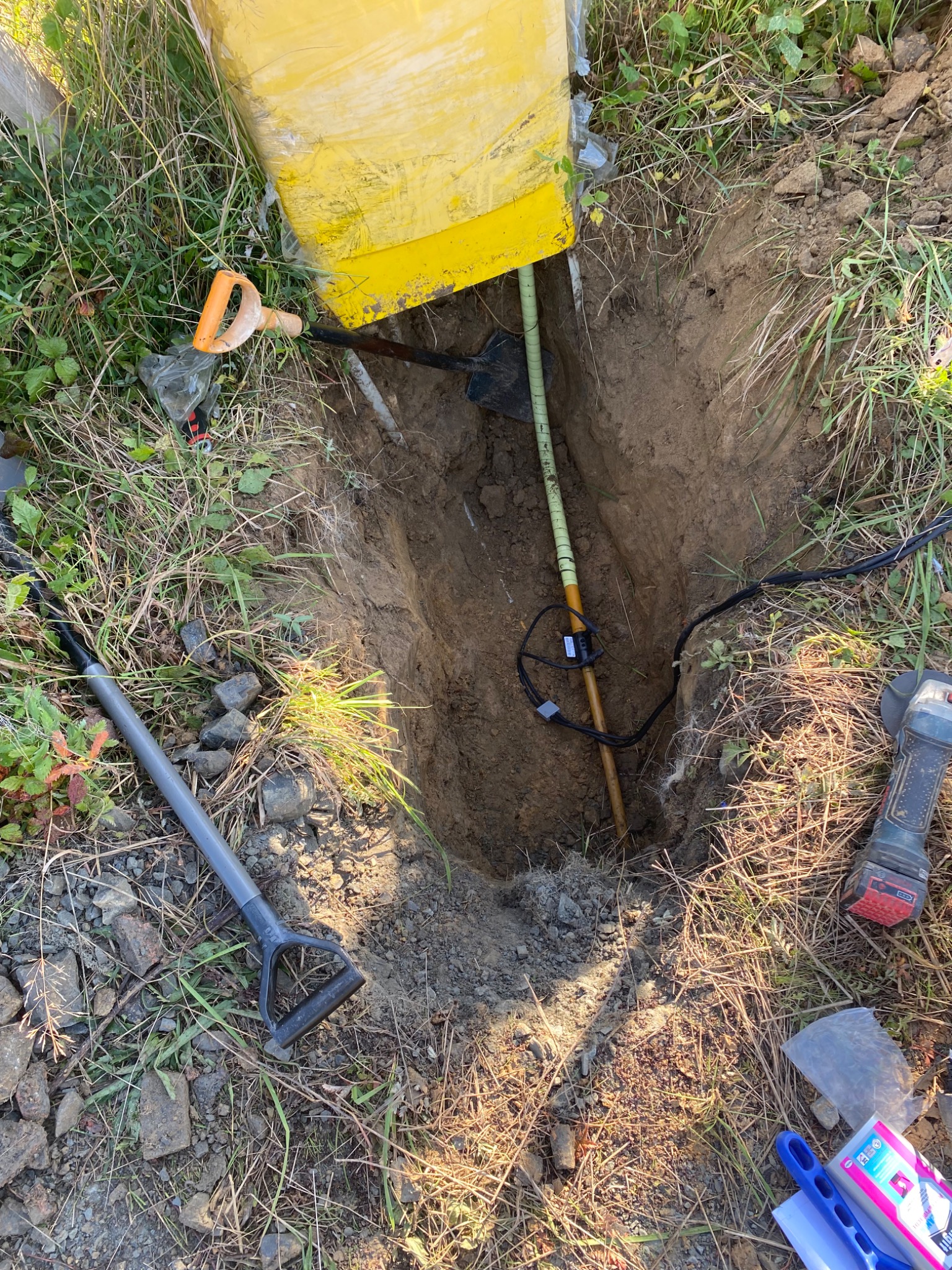 Wykop w ziemi z wystającą rurą z podłączonymi przewodami, obok leżą łopaty i szlifierka kątowa.
