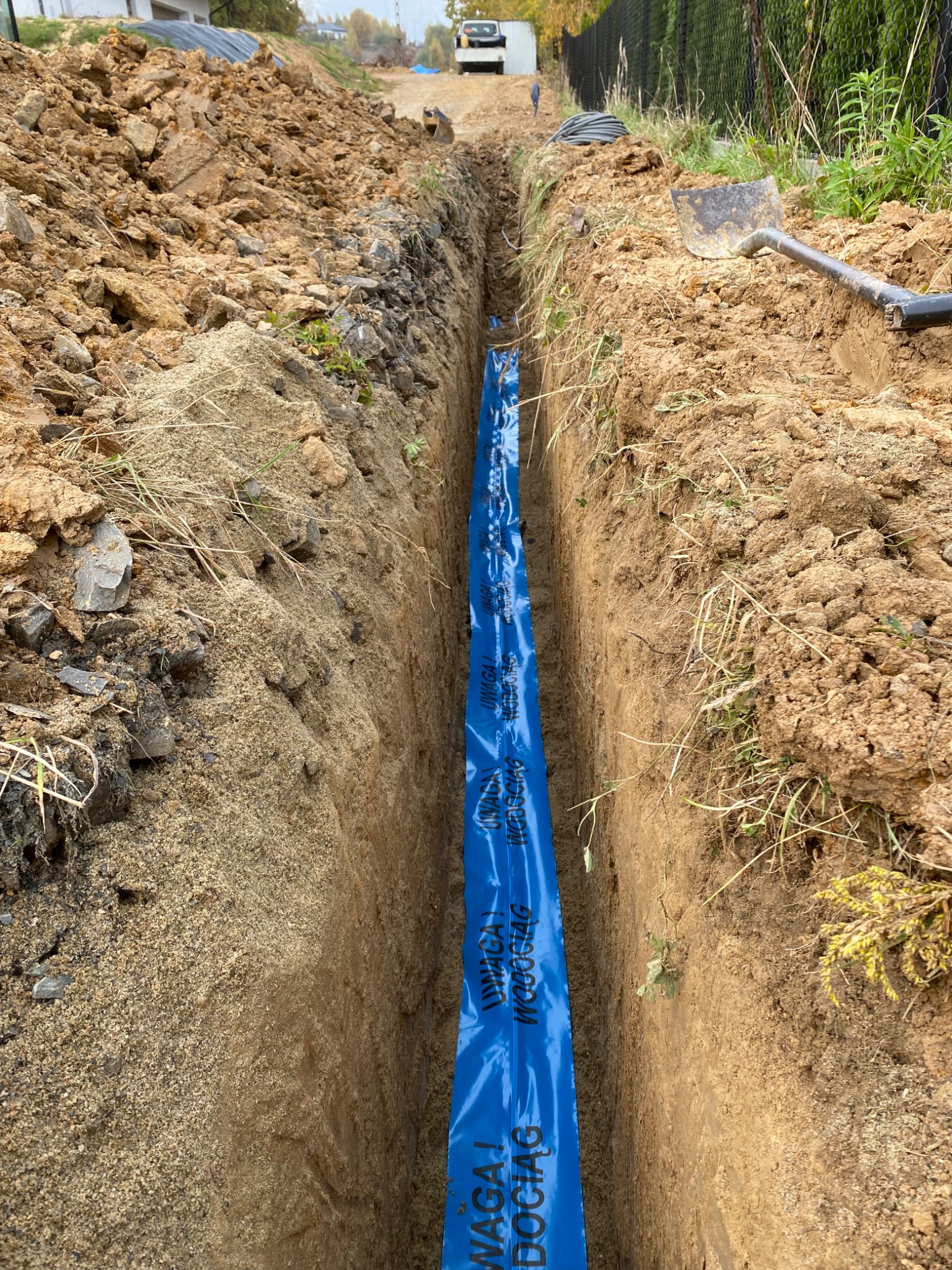Wykop w ziemi z niebieską folią ostrzegawczą 'UWAGA! WODOCIĄG' ułożoną na dnie, widoczna łopata i zwój kabli na krawędzi wykopu, w tle zabudowa jednorodzinna i sprzęt budowlany.