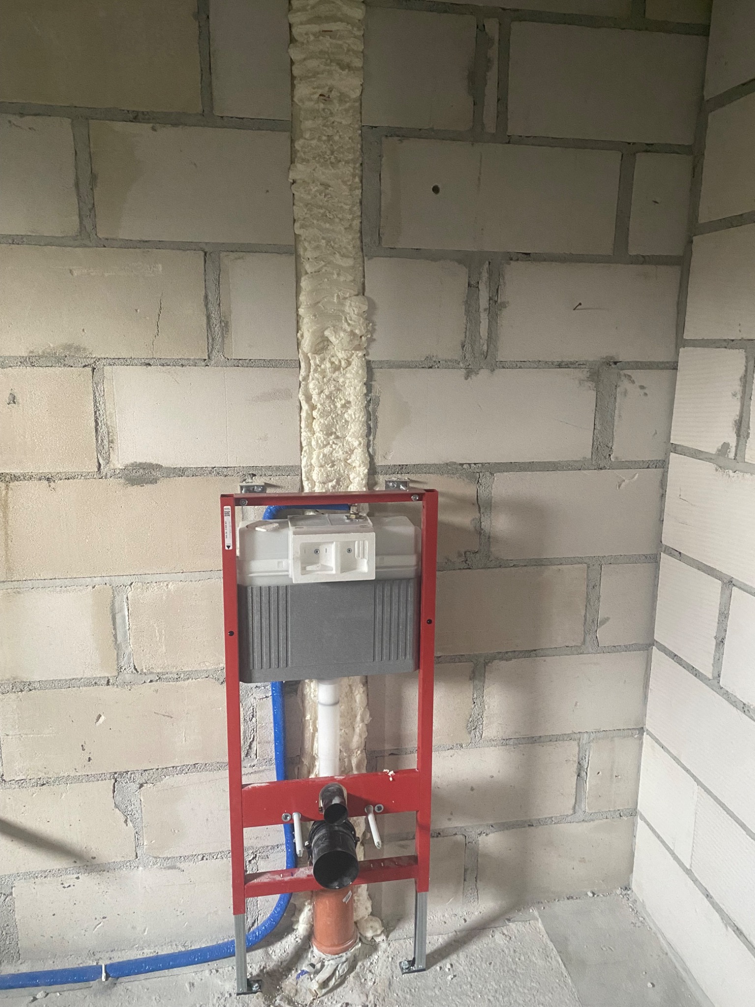 Stelaż podtynkowy WC zamontowany na ścianie z betonu komórkowego, rura kanalizacyjna z uszczelnieniem, niebieska rura doprowadzająca wodę, szczelina w ścianie wypełniona pianką montażową.