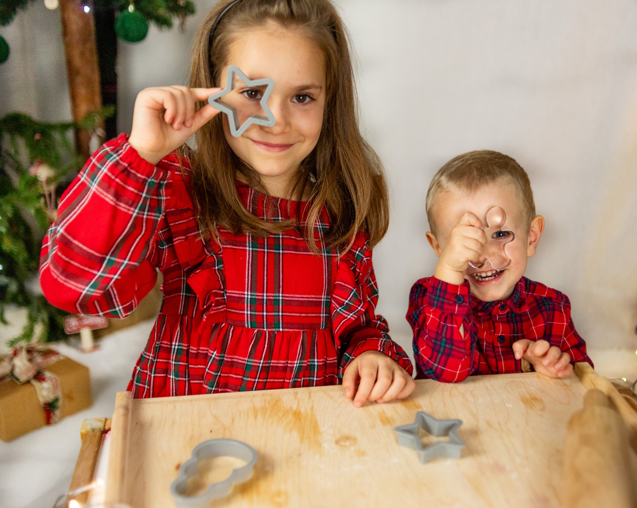 Dwoje uśmiechniętych dzieci w czerwonych, kraciastych ubraniach bawi się foremkami do ciastek w kształcie gwiazdy i ludzika na tle dekoracji świątecznych.