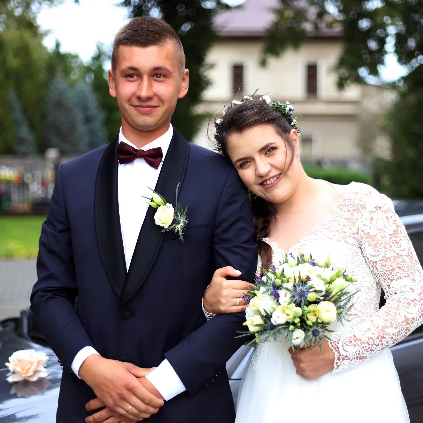 Uśmiechnięta para młoda w strojach ślubnych: on w granatowym smokingu z bordową muchą, ona w białej sukni z koronkowymi rękawami i wiankiem na głowie, trzyma bukiet z białych i fioletowych kwiatów,...