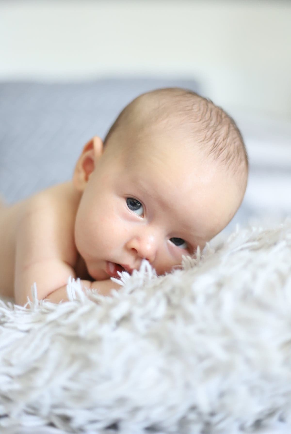 Niemowlę leżące na puszystym, białym kocyku, z wyciągniętym językiem i niebieskimi oczami, patrzy w obiektyw.