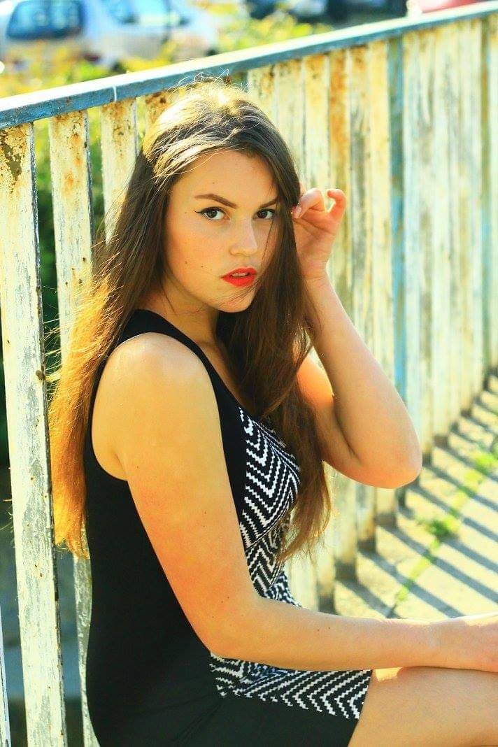 Portret młodej kobiety z długimi brązowymi włosami, w czarnej sukience z geometrycznym wzorem, pozującej na tle zardzewiałej, pomalowanej na biało barierki.