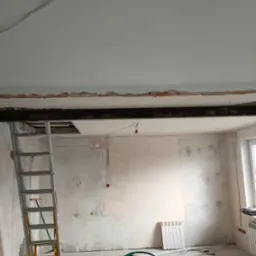 Wnętrze w trakcie remontu: surowe ściany, odsłonięta belka stropowa, drabina oparta o ścianę, grzejnik i narzędzia na podłodze, widoczne ślady po instalacjach elektrycznych.