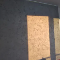 Ściana pokryta dekoracyjną szarą strukturą, oświetlona popołudniowym słońcem tworzącym wyraźny cień, z widoczną taśmą malarską przy suficie.