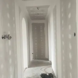 Korytarz w trakcie prac wykończeniowych: ściany pokryte płytami gipsowo-kartonowymi, widoczne otwory po wkrętach i nierówności, surowy beton na podłodze, głośnik leżący na podłodze, instalacja...