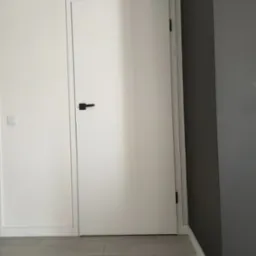 Minimalistyczne wnętrze: białe drzwi z czarną klamką na tle szarej ściany, jasna podłoga z paneli.