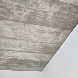 Patryk Furlepa Budownictwo remontowo wykończeniowe - Wykończony sufit z efektem betonu architektonicznego, widoczny narożnik pomieszczenia z białymi ścianami. Realizacja wykończenia wnętrza z bliska.
