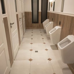 Patryk Furlepa Budownictwo remontowo wykończeniowe - Publiczna toaleta: rzędy białych pisuarów na tle drewnianej boazerii i białych drzwi kabin. Wzór na podłodze z płytek w romby w odcieniach beżu.