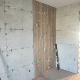 Patryk Furlepa Budownictwo remontowo wykończeniowe - Układanie płytek w łazience: szare płytki imitujące beton i drewnopodobne, pionowe panele z użyciem systemu poziomowania. Widoczna poziomica i laserowa linia.