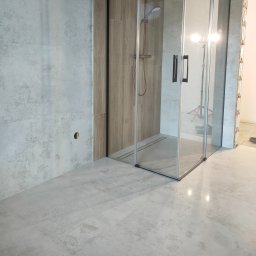 Patryk Furlepa Budownictwo remontowo wykończeniowe - Nowoczesna łazienka w trakcie wykańczania: szklana kabina prysznicowa, szare płytki na ścianach i podłodze, widoczne elementy instalacji. Oświetlenie robocze wewnątrz kabiny.