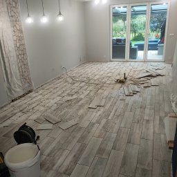 Patryk Furlepa Budownictwo remontowo wykończeniowe - Układanie podłogi z płytek imitujących drewno w jasnym pomieszczeniu z dużym oknem tarasowym. Widoczne narzędzia i resztki kleju. Etap prac w trakcie realizacji.