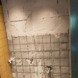 Patryk Furlepa Budownictwo remontowo wykończeniowe - Fragment ściany łazienki w trakcie skuwania starych płytek, widoczne pozostałości kleju i fragmenty płytek na podłodze. Żółte płytki w rogu.