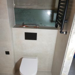 Patryk Furlepa Budownictwo remontowo wykończeniowe - Wykończona łazienka z beżowymi płytkami, wiszącą toaletą i czarnym grzejnikiem drabinkowym. Widoczne niedokończone fragmenty ściany.