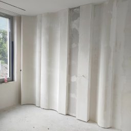 Patryk Furlepa Budownictwo remontowo wykończeniowe - Narożnik pokoju z falistą ścianą z płyt gipsowo-kartonowych w trakcie szpachlowania. Widoczne kable i niedoskonałości powierzchni. Okno z widokiem na zieleń.