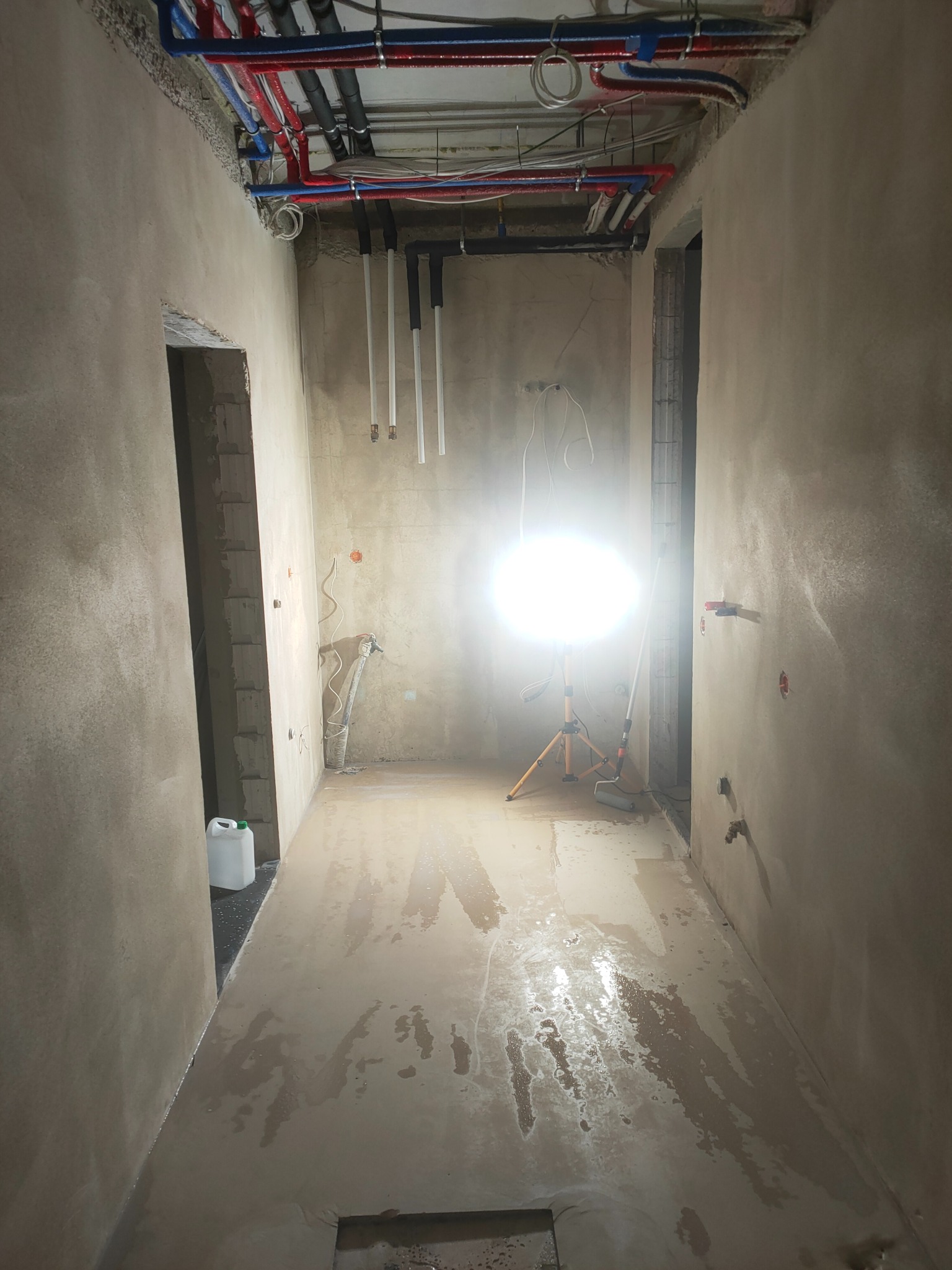 Wnętrze w trakcie prac wykończeniowych: ściany pokryte świeżą gładzią, widoczne rury instalacji pod sufitem, przenośna lampa budowlana oświetlająca pomieszczenie, mokra podłoga i wiadro...
