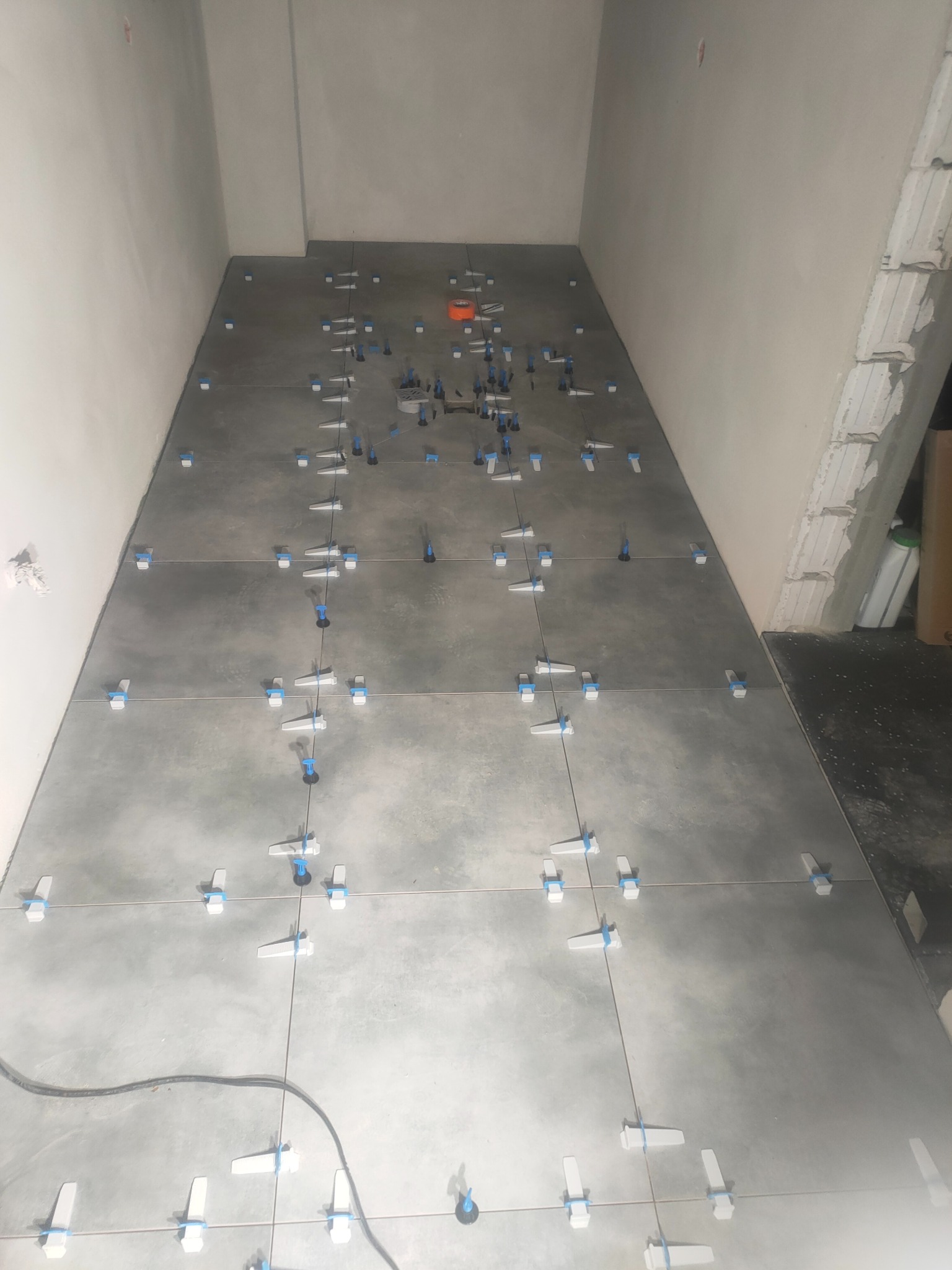 Układanie szarych płytek podłogowych z użyciem klipsów poziomujących w trakcie prac remontowych. Widoczny przewód elektryczny na podłodze.