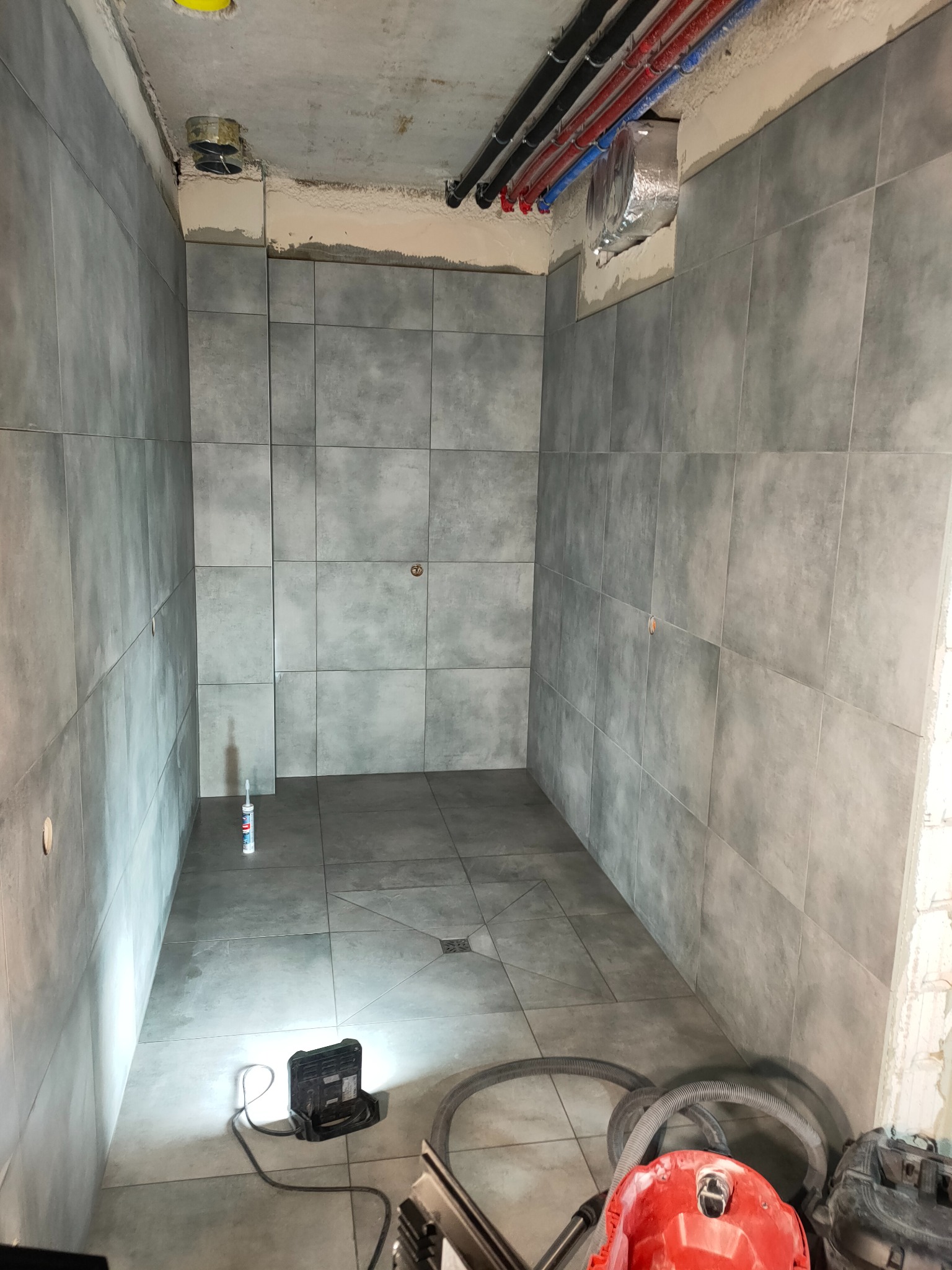 Wnętrze łazienki w trakcie remontu, ściany i podłoga wyłożone szarymi płytkami, widoczny silikon uszczelniający, lampa budowlana i odkurzacz przemysłowy.