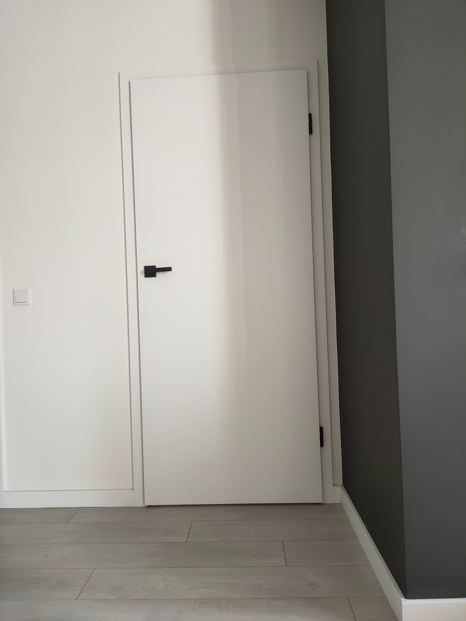 Minimalistyczne wnętrze: białe drzwi z czarną klamką na tle szarej ściany, jasna podłoga z paneli.