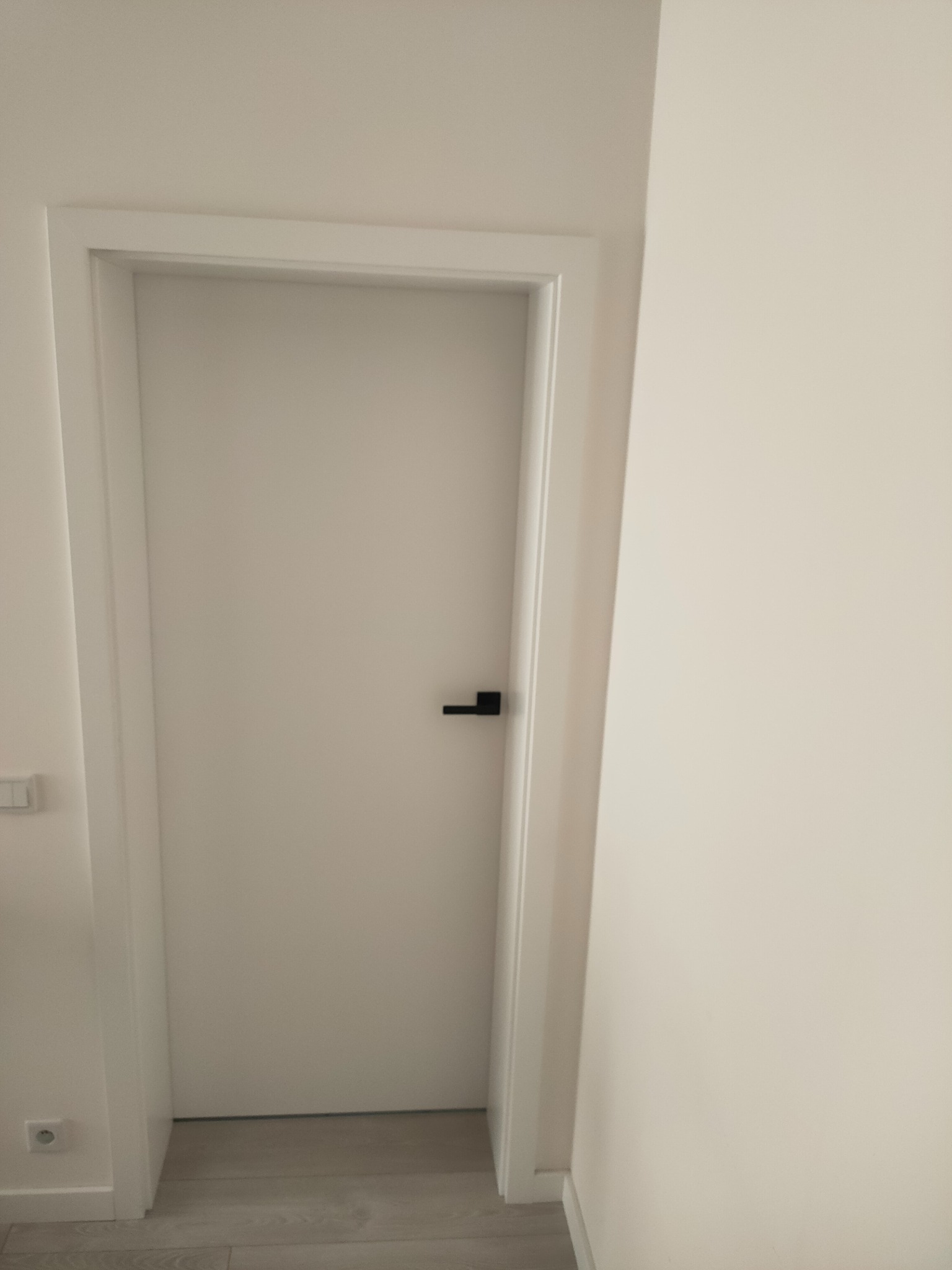 Białe drzwi wewnętrzne z czarną klamką na tle jasnej ściany. Minimalistyczny design, widoczny fragment podłogi z paneli.