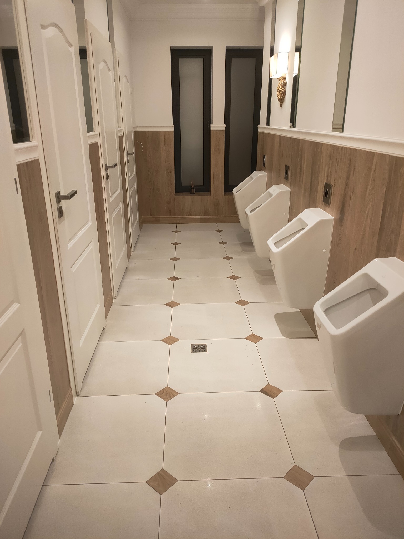 Publiczna toaleta: rzędy białych pisuarów na tle drewnianej boazerii i białych drzwi kabin. Wzór na podłodze z płytek w romby w odcieniach beżu.