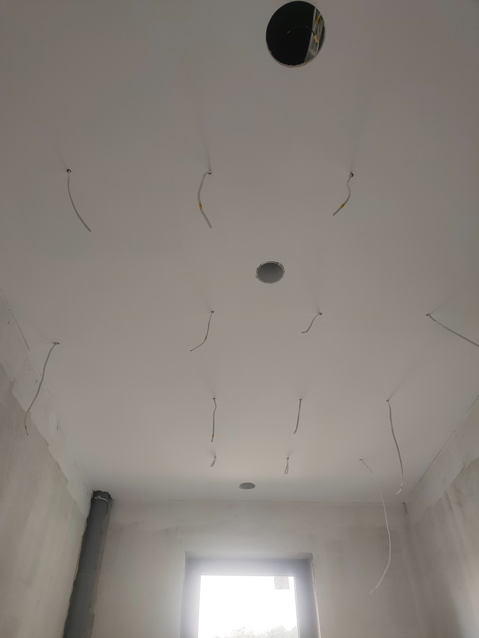 Biały sufit z wystającymi przewodami elektrycznymi i otworami na oświetlenie, w trakcie prac wykończeniowych. Widoczne ślady po szpachlowaniu, okno z jasnym światłem.