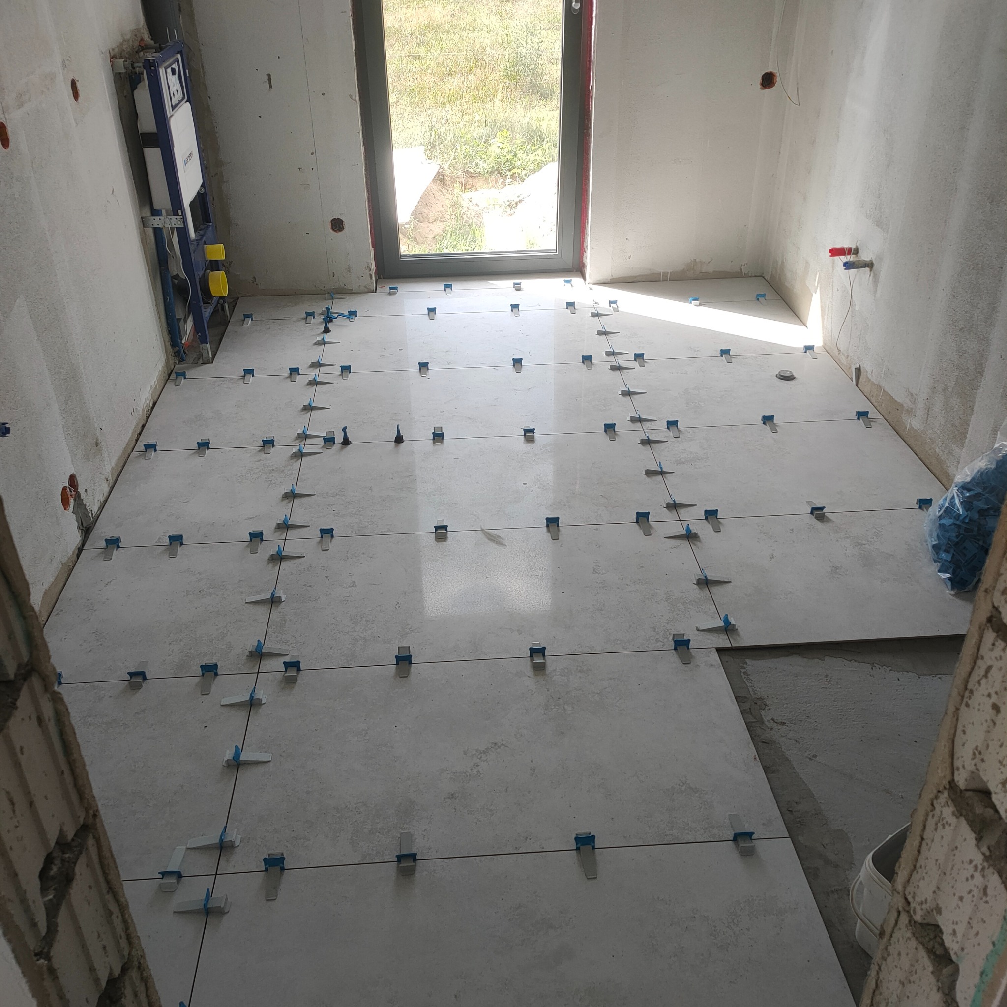 Jasnoszare płytki podłogowe w trakcie układania, z użyciem niebieskich klipsów poziomujących. Widoczna instalacja podtynkowa WC i okno z widokiem na zieleń.