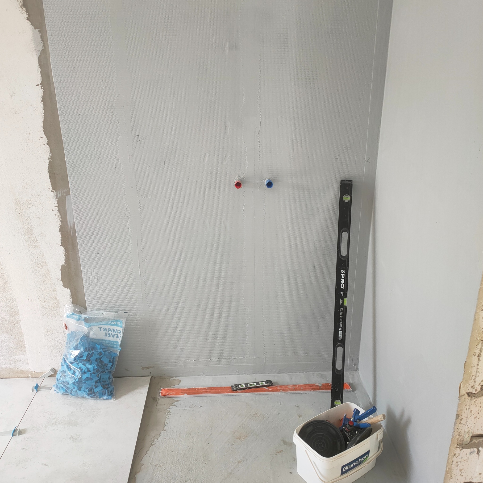 Przygotowanie podłoża pod płytki w łazience: ściana z siatką, poziomica, narzędzia w wiadrze, paczka z niebieskimi klipsami poziomującymi.