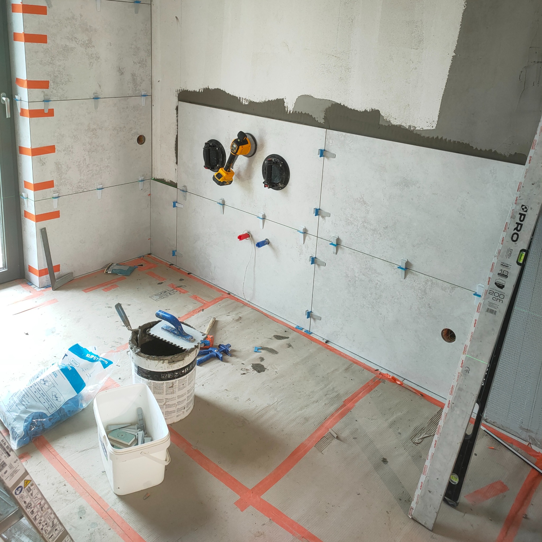 Ściana łazienki w trakcie układania płytek, widoczne przyssawki, poziomica laserowa, krzyżyki dystansowe. Podłoga oznaczona taśmą.