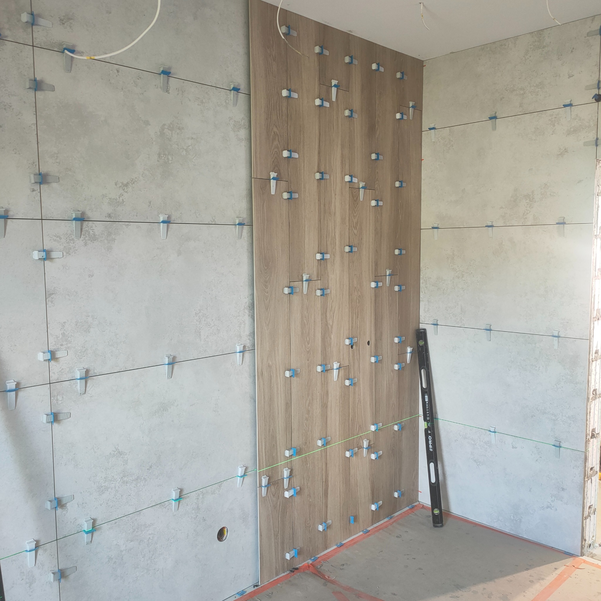 Układanie płytek w łazience: szare płytki imitujące beton i drewnopodobne, pionowe panele z użyciem systemu poziomowania. Widoczna poziomica i laserowa linia.