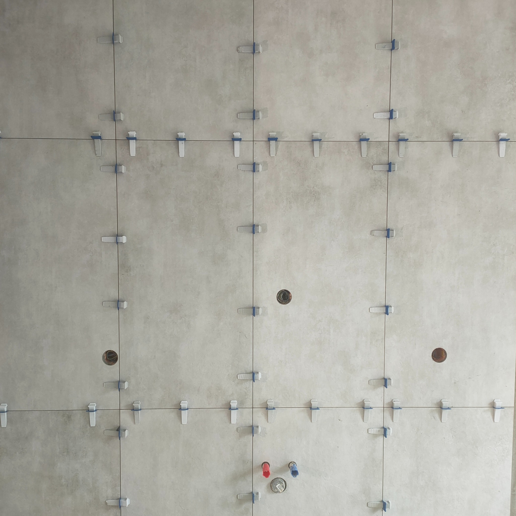 Ściana z jasnych płytek, wyrównywana za pomocą klipsów i krzyżyków. Widoczne otwory na instalacje. Etap prac wykończeniowych w łazience.