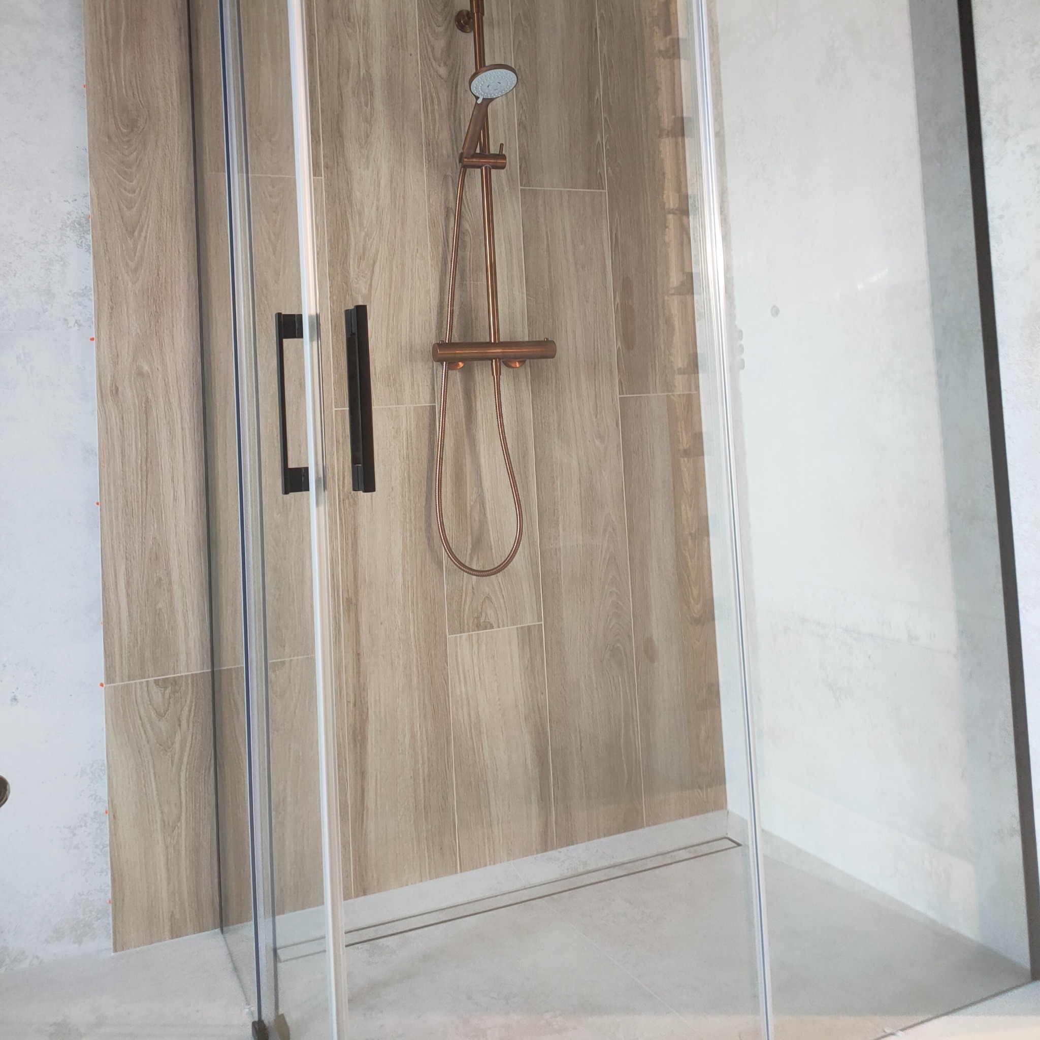 Kabina prysznicowa z brązową armaturą na tle ściany wyłożonej płytkami imitującymi drewno. Minimalistyczny design, nowoczesna łazienka po remoncie.