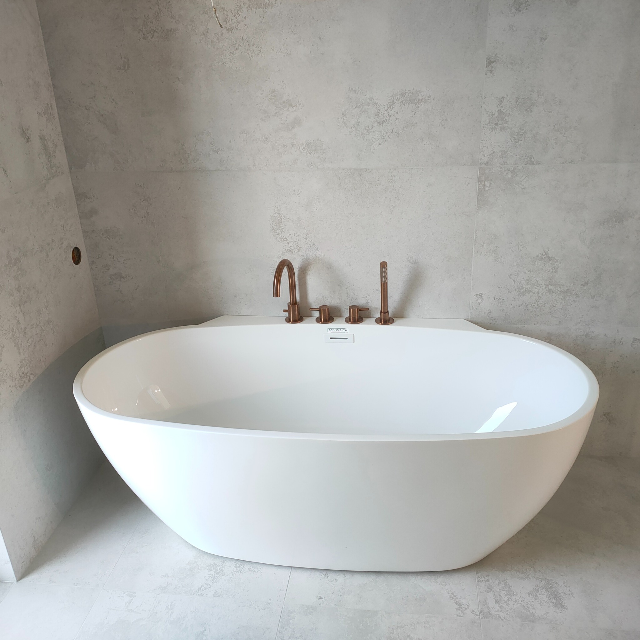 Nowoczesna, wolnostojąca wanna w jasnej łazience z szarymi płytkami imitującymi beton i miedzianą armaturą. Minimalistyczny design, wysoka jakość wykończenia.