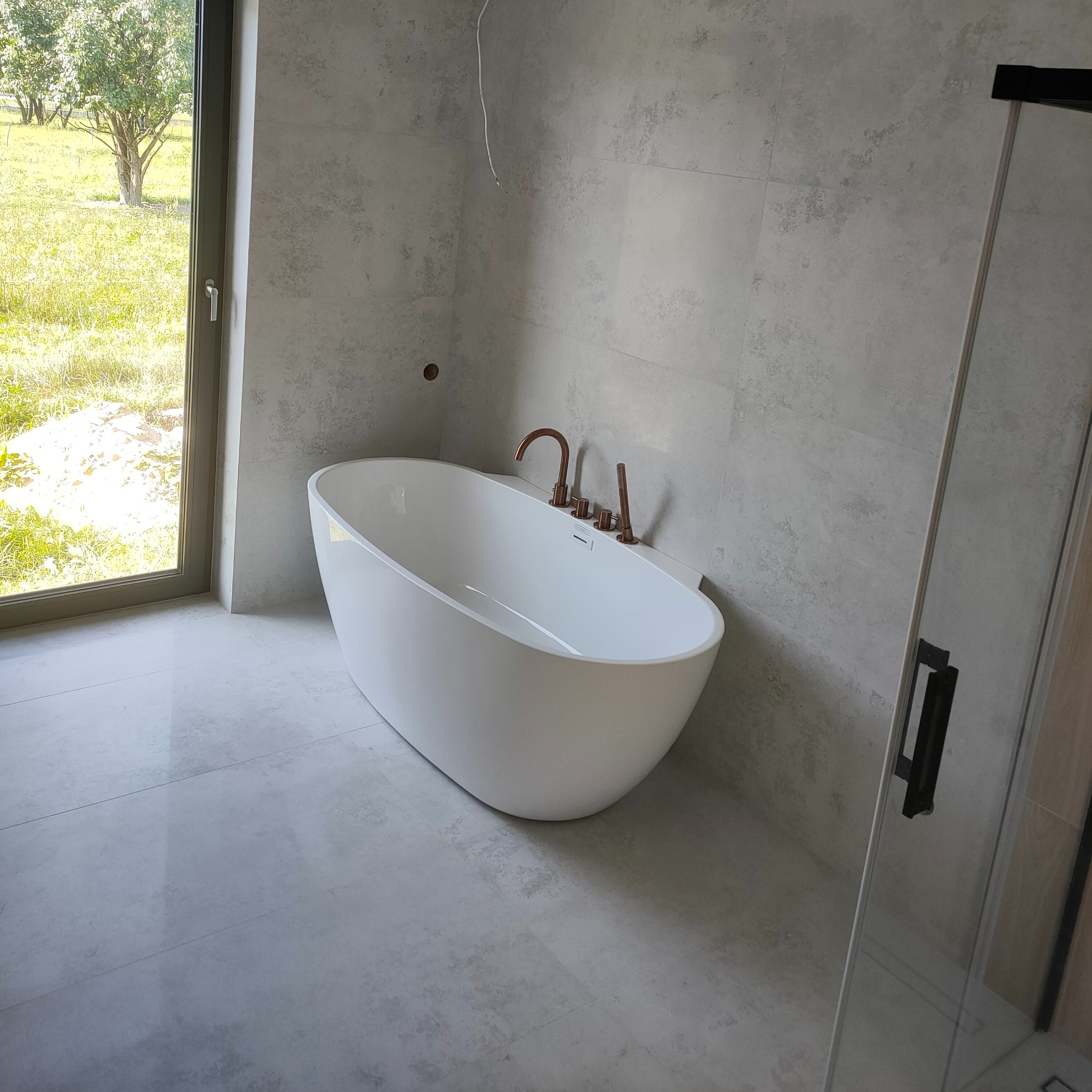 Nowoczesna łazienka z wolnostojącą białą wanną, szarą glazurą i miedzianą armaturą. Okno z widokiem na zieleń. Kabina prysznicowa w tle.