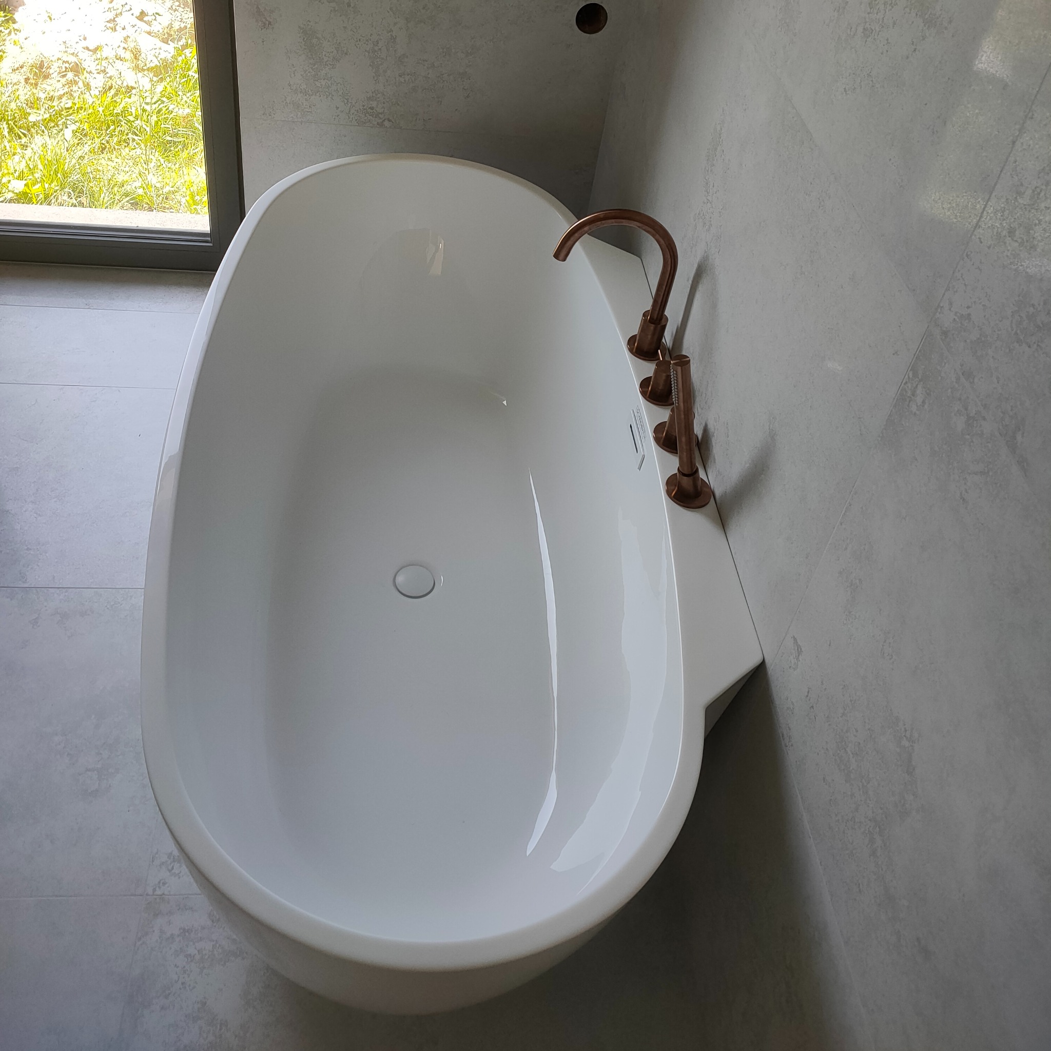 Nowoczesna, wolnostojąca wanna z miedzianą armaturą na tle ściany wyłożonej szarą, strukturalną płytką w łazience z oknem. Minimalistyczny design.