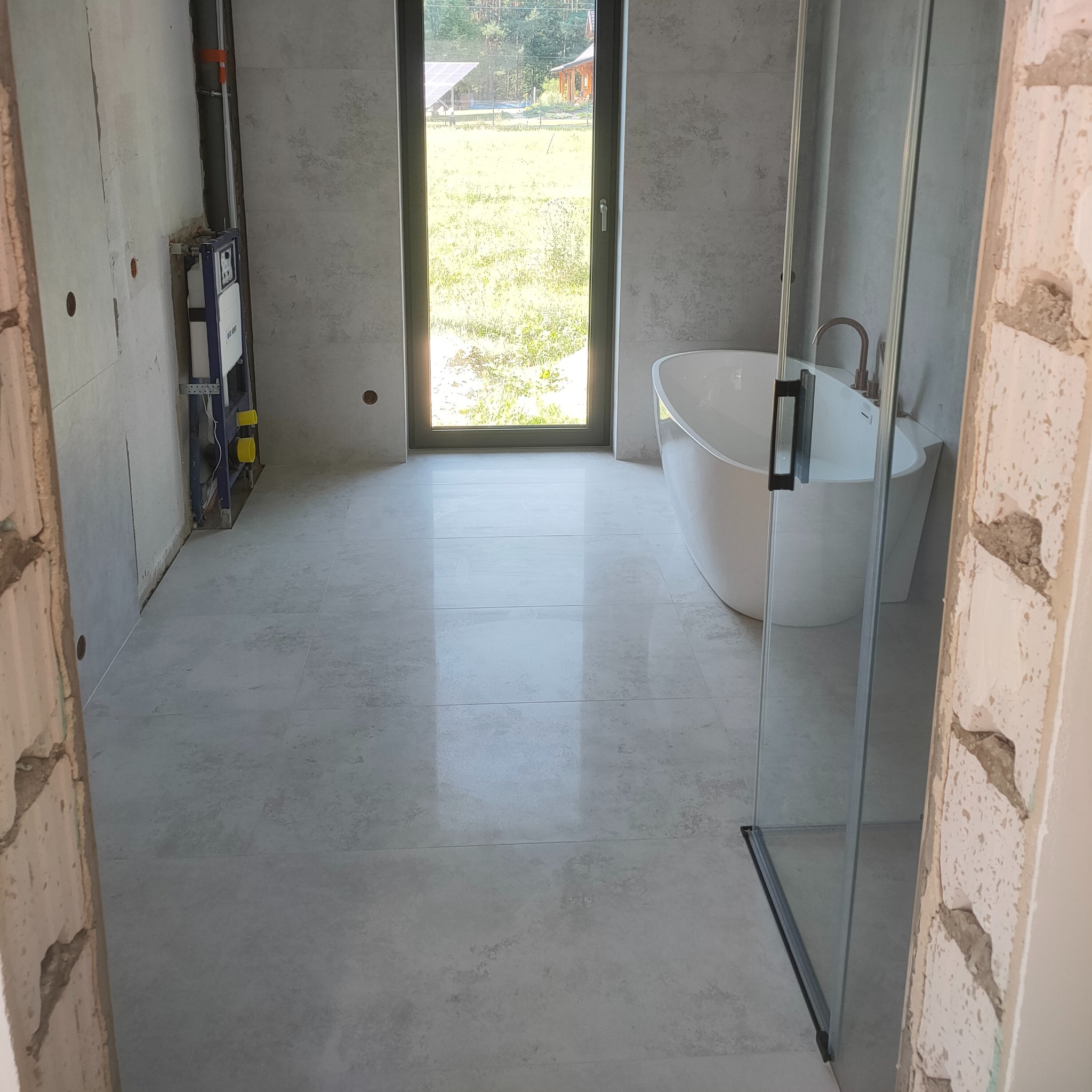 Łazienka w trakcie remontu: ułożone szare płytki podłogowe, zamontowana instalacja podtynkowa WC, widoczna wanna i kabina prysznicowa. Tło stanowi okno z widokiem na zieleń.