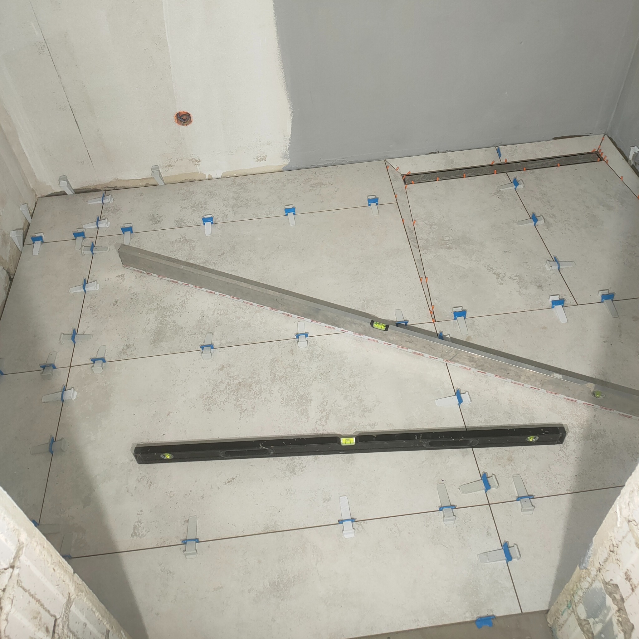 Układanie płytek podłogowych z użyciem poziomnic i klipsów poziomujących w jasnym wnętrzu łazienki w trakcie remontu. Widoczne fugi i fragment ściany.