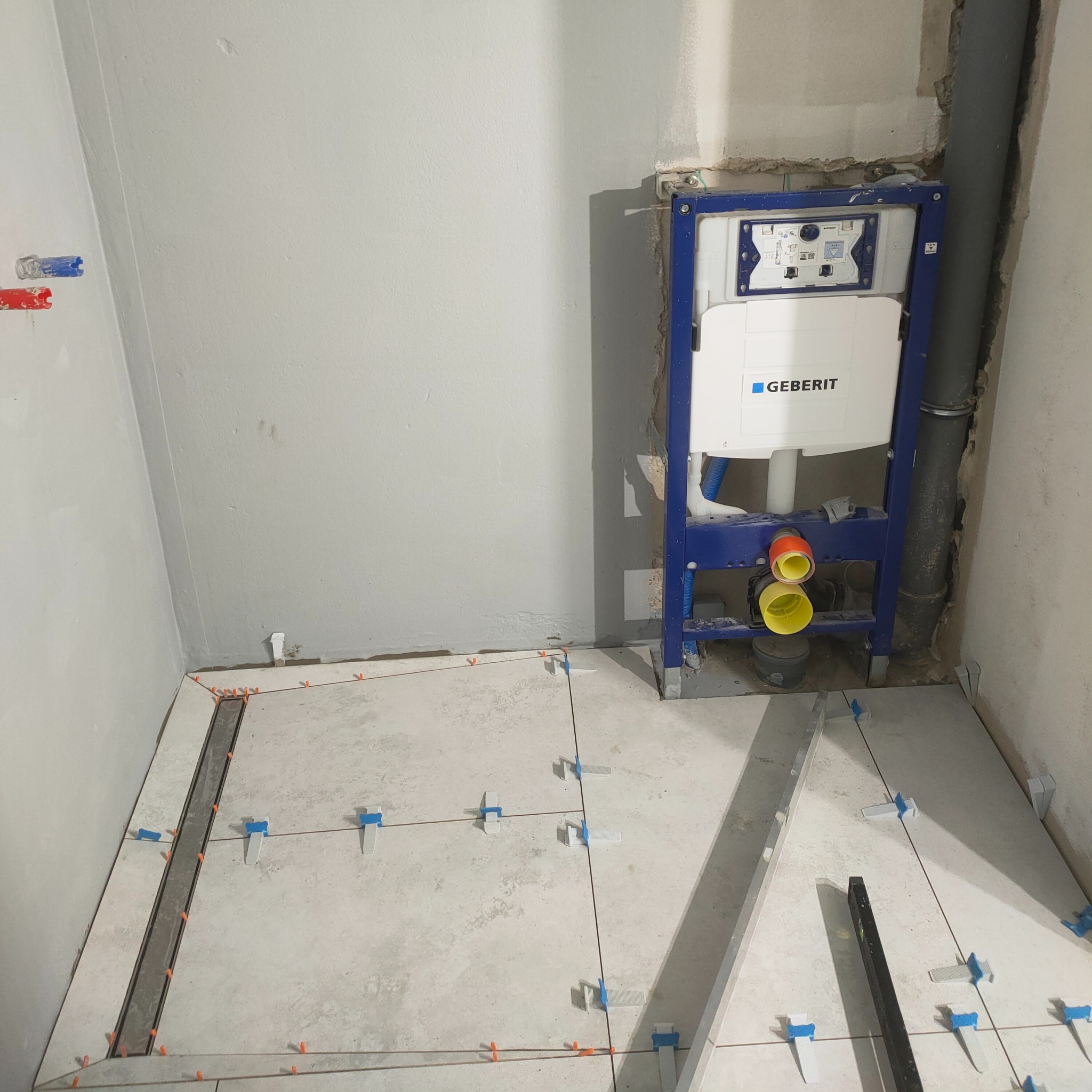 Montaż stelaża podtynkowego Geberit i układanie jasnych płytek podłogowych z użyciem poziomicy i krzyżyków w łazience w trakcie remontu.