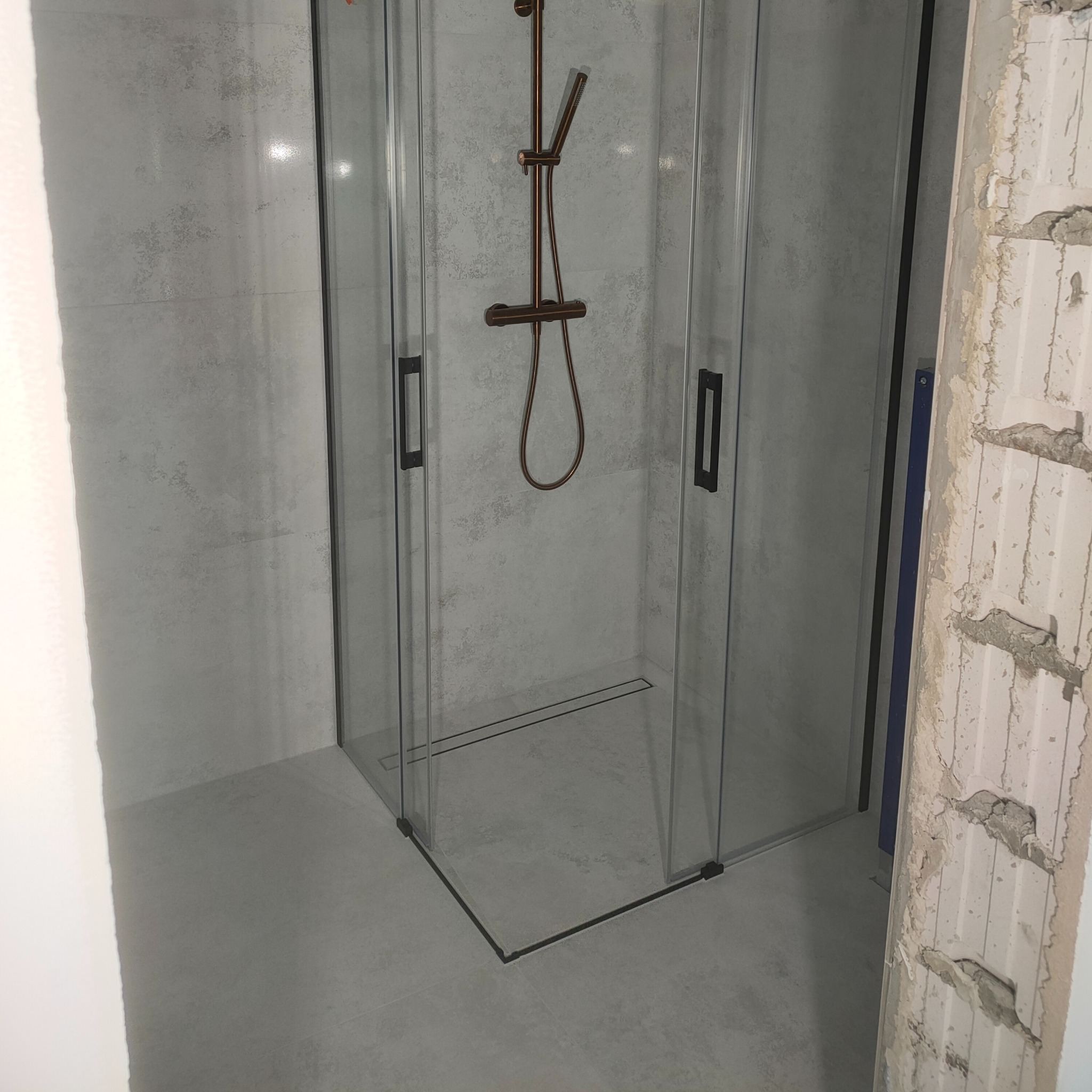 Nowoczesna kabina prysznicowa z brązową armaturą na tle szarych płytek. Fragment ściany w stanie surowym. Widoczny odpływ liniowy.