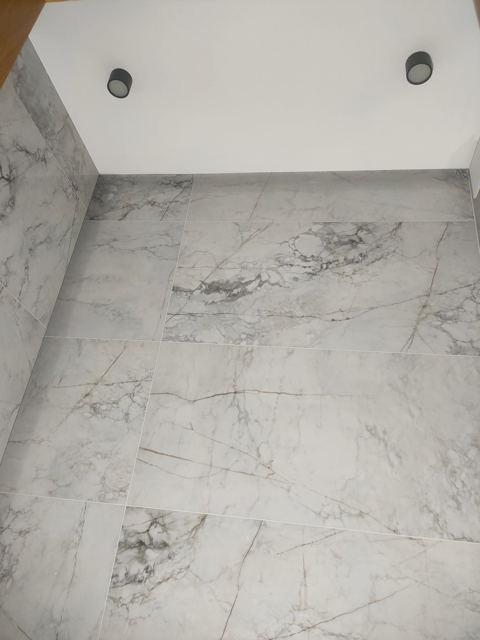 Wykończona łazienka z szarymi płytkami imitującymi marmur na ścianach i podłodze, widoczne dwa czarne punkty oświetleniowe w suficie.