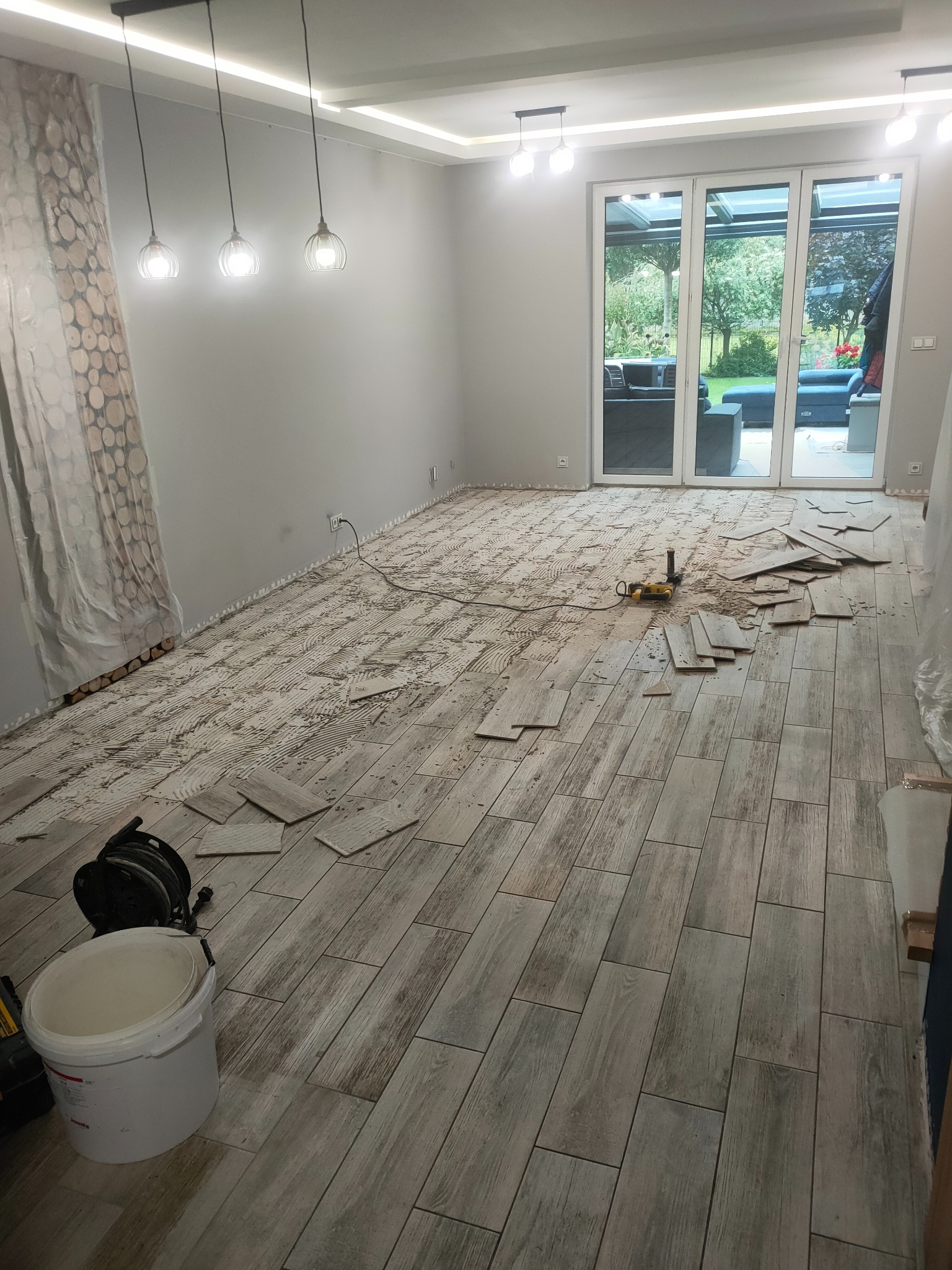 Układanie podłogi z płytek imitujących drewno w jasnym pomieszczeniu z dużym oknem tarasowym. Widoczne narzędzia i resztki kleju. Etap prac w trakcie realizacji.