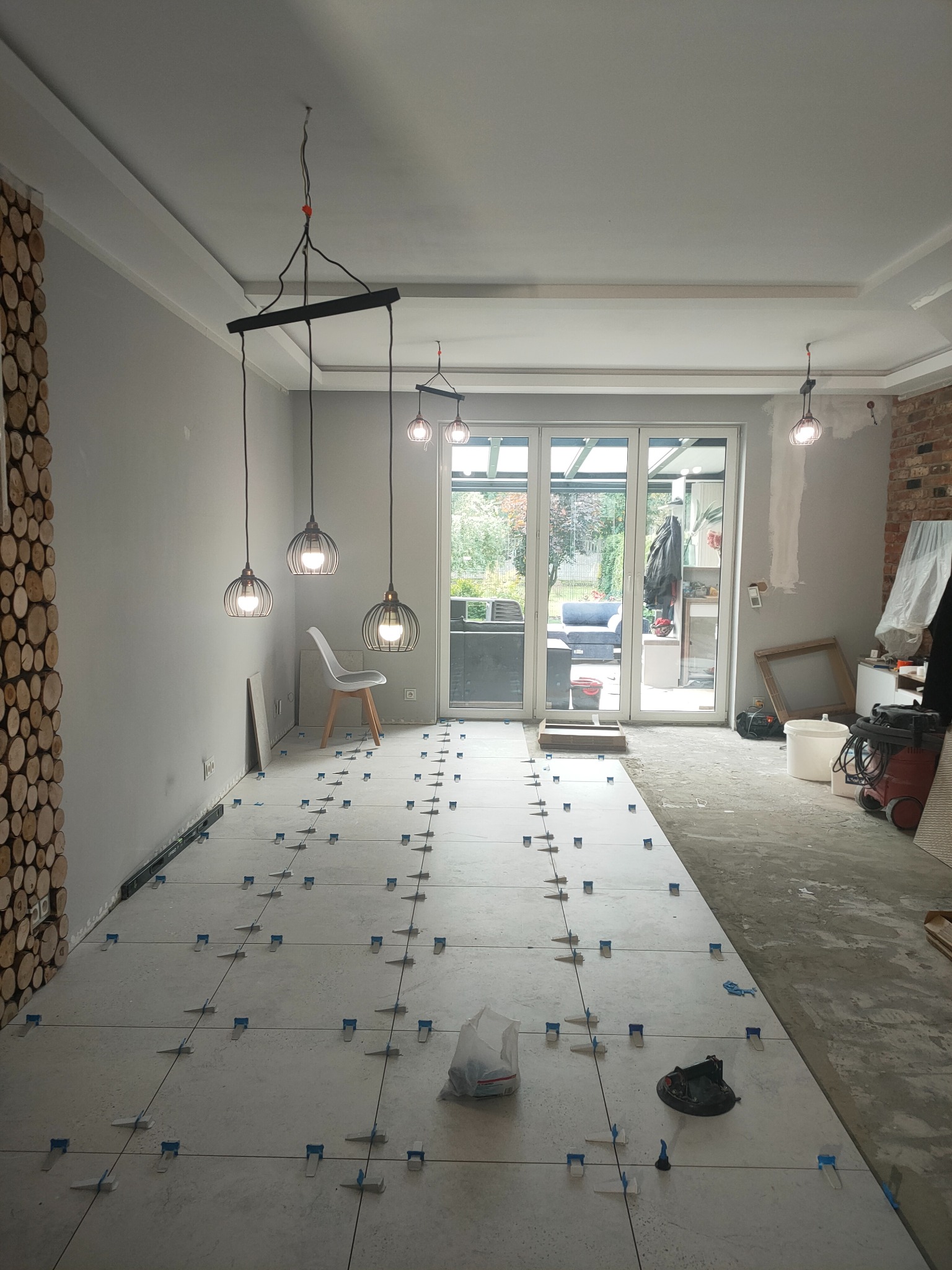 Układanie jasnych płytek podłogowych z użyciem systemu poziomowania w Łodzi. Widoczne narzędzia i fragmenty surowego wykończenia ścian.