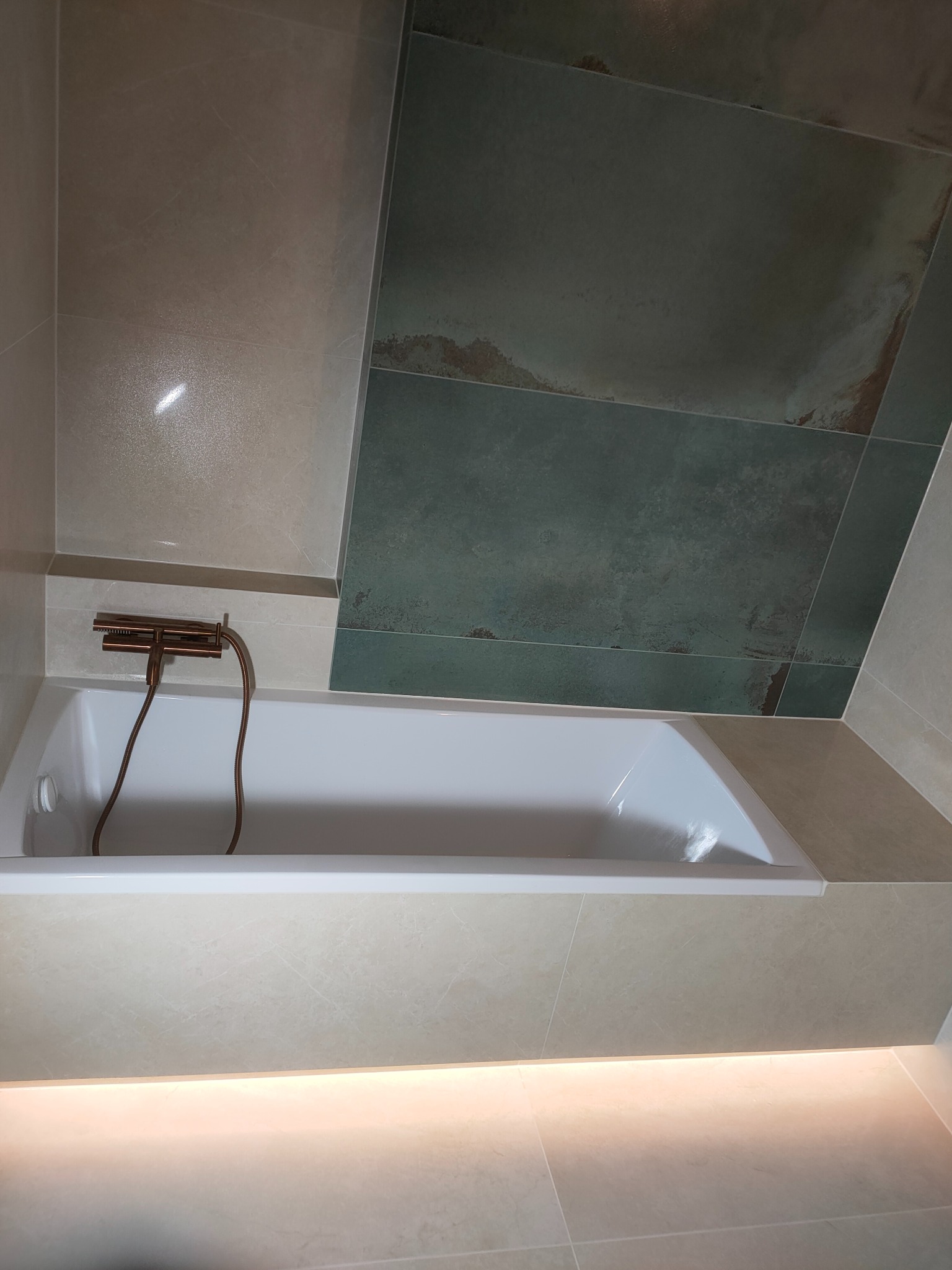 Nowoczesna łazienka z wanną, beżowymi płytkami i akcentem zieleni na ścianie. Miedziana armatura dodaje elegancji. Podświetlenie LED podkreśla minimalistyczny design.