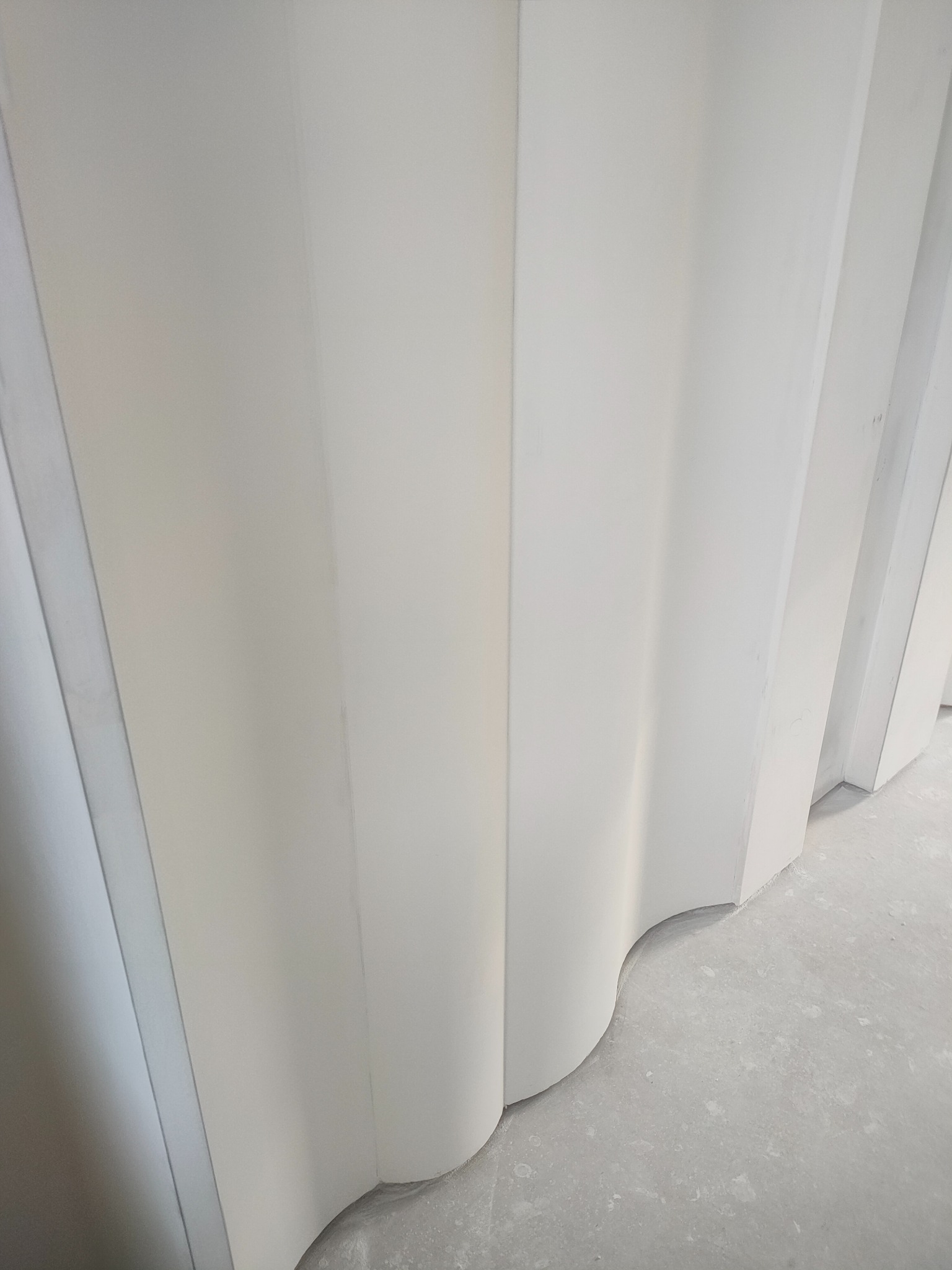 Białe, faliste panele ścienne z zaokrąglonymi krawędziami, ułożone pionowo, widoczne z dołu pod kątem, na szarym, surowym podłożu. Minimalistyczny design.