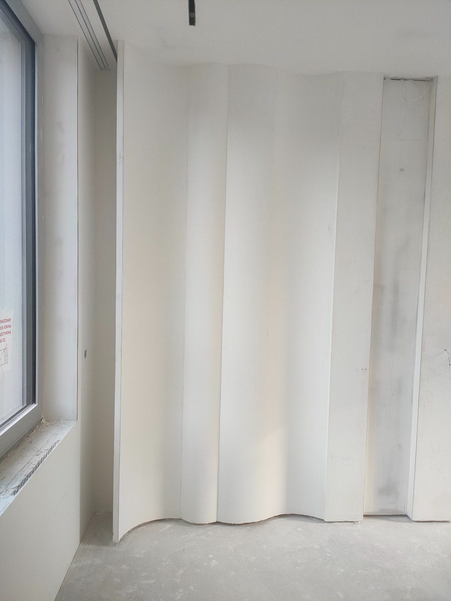 Biała, falista ścianka działowa z płyty gipsowej w surowym wnętrzu, widoczna przy oknie. Szare, betonowe podłoże. Minimalistyczny design, surowy stan wykończenia.