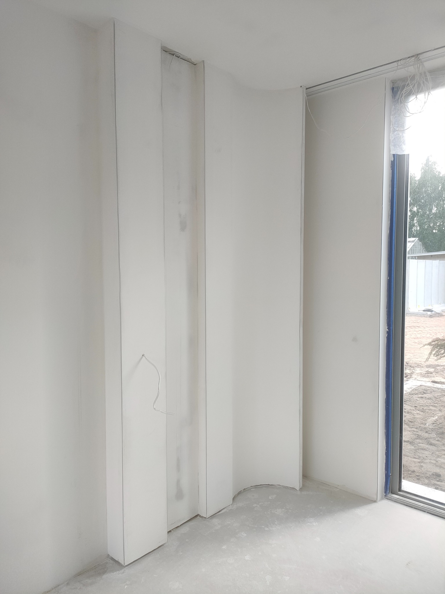 Białe ściany z płyt kartonowo-gipsowych w surowym wnętrzu, widoczne kable i okno z widokiem na zewnątrz. Minimalistyczny kadr, surowy stan wykończenia.