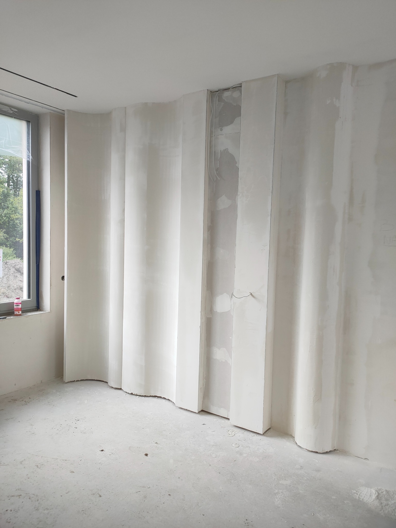 Narożnik pokoju z falistą ścianą z płyt gipsowo-kartonowych w trakcie szpachlowania. Widoczne kable i niedoskonałości powierzchni. Okno z widokiem na zieleń.