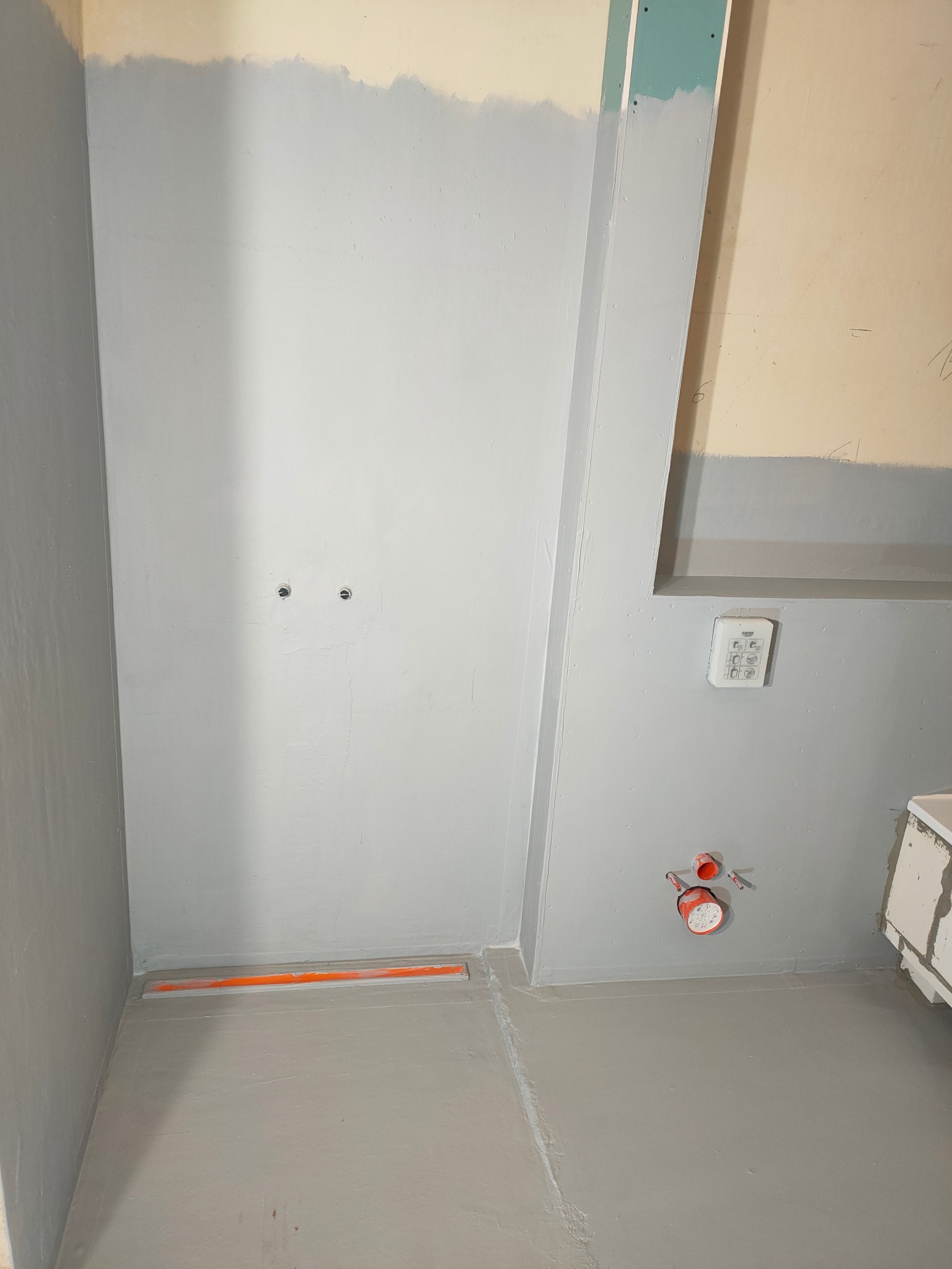 Szara łazienka w trakcie remontu: widoczny odpływ liniowy, przygotowane ściany pod glazurę, instalacja wodna. Etap prac przed wykończeniem.